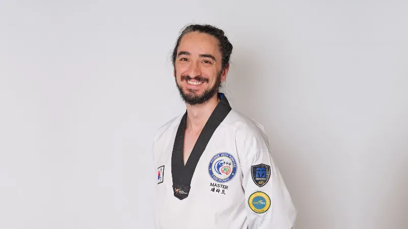 Taekwondo instructor wearing a white uniform and black belt standing against a plain background.