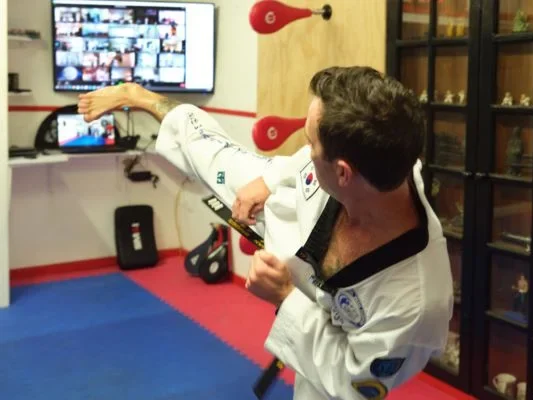 Taekwondo instructor practising a kick while following an online martial arts class on a screen.