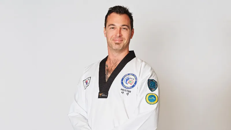 Taekwondo instructor wearing a white uniform and black belt standing against a plain background.