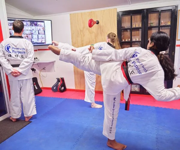 Taekwondo students practising kicking techniques while following an online martial arts class on a screen.