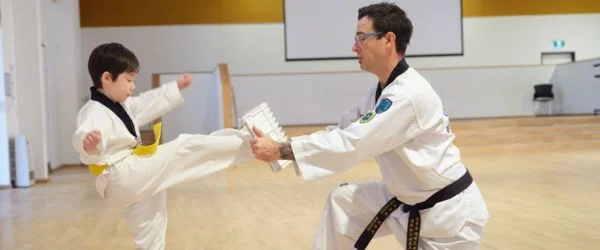 Young Taekwondo student practising a front kick while an instructor holds a target pad during class.