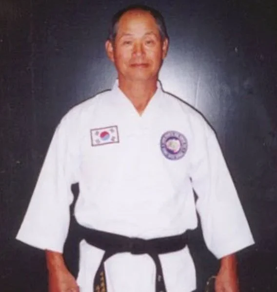Senior Taekwondo master wearing a white uniform and black belt standing against a dark background.