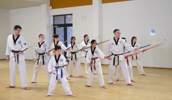 Group of students and instructors in white taekwondo uniforms practising staff techniques in a martial arts class.