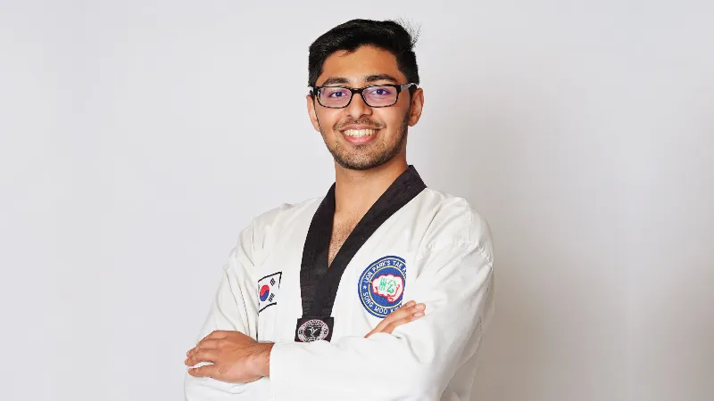 Taekwondo instructor wearing a white uniform and black belt standing with arms crossed against a plain background.