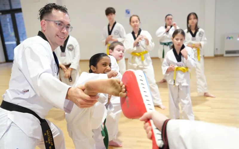 Martial arts class: instructor shields a student’s kick with a padded target while others watch in white uniforms.