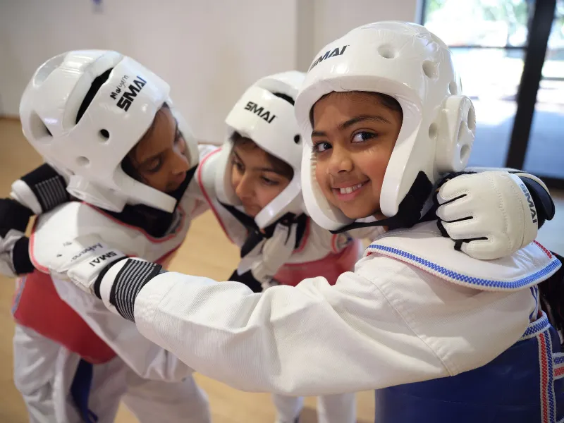 Three smiling children in white protective helmets and gis train together in a gym, arms linked in a team pose for taekwondo practice.