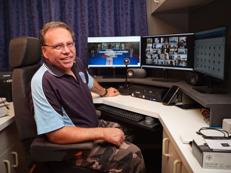 Smiling man in a polo shirt sits at a desk with multiple monitors during a video conference setup.