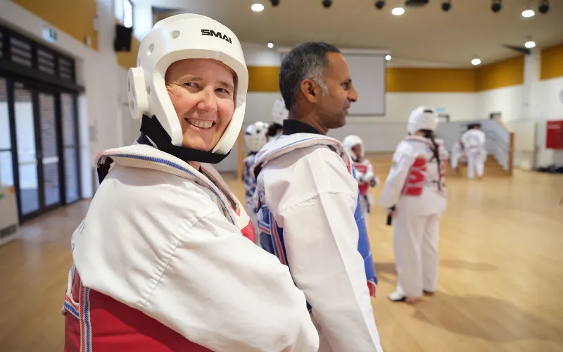 Smiling woman in a white taekwondo uniform and helmet, preparing for training in a dojo with others in the background.